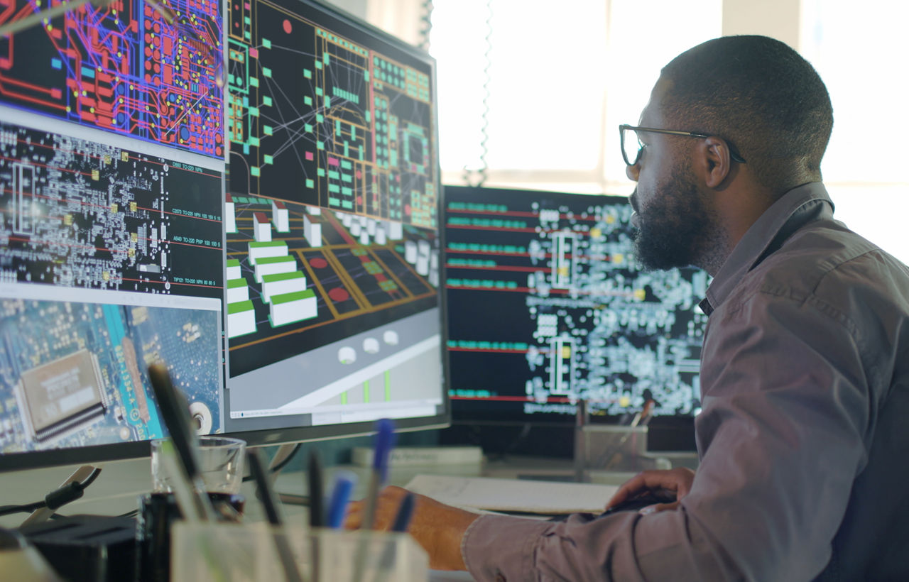 Stock image of an Afro-Caribbean male designing electronic circuit boards ( PCBs).
Heâ  s sitting at a desk with a large computer screen displaying a variety of apps & PCB illustrations.